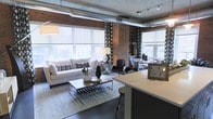 This single-floor urban loft masterfully combines raw industrial aesthetics with contemporary sophistication. Exposed brick walls and concrete ceilings with visible ductwork and track lighting define the architectural character, while large windows fitted with roller shades flood the open-plan interior with abundant natural light and offer sweeping city views.

The open living area is anchored by a plush white sofa, a glass-and-metal coffee table, and a textured area rug that adds warmth to the space. Bold patterned curtains frame the windows, providing a striking contrast to the otherwise neutral palette. A sleek kitchen island with bar seating transitions seamlessly into the living zone, reinforcing the loft's open and connected layout.

The dining area sits adjacent to the living space, furnished with dark chairs arranged around a round table, set against a backdrop of brick and large windows. Throughout the space, carefully curated décor — including statement lamps, mirrored accents, and sculptural objects — adds layers of texture and visual interest.

The overall ambiance is one of modern urban living: industrial in structure, yet refined and warm in execution. The interplay of natural light, exposed materials, and thoughtfully selected furnishings creates an inviting and dynamic environment.