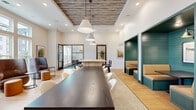 Located at 713 Adelynn Ln in Greensboro, NC, this single-floor communal space combines modern design with warm, inviting finishes across a well-appointed layout. The dining area serves as a central hub, anchored by a long, dark wood communal table paired with upholstered seating and black spindle-back bar stools along a fluted-panel counter. Overhead, a striking reclaimed wood ceiling inset adds rustic character, complemented by pendant lights with brass accents. A row of teal shiplap-lined booth nooks lines one wall, offering intimate seating areas ideal for smaller gatherings or focused conversations. The living room area features soaring ceilings with expansive windows that flood the space with natural light and offer views of the surrounding community. Plush lounge chairs and a teal built-in shelving unit add color and function to this relaxed zone. A private meeting room, accessible via glass-panel doors, includes a round marble-top table, leather rolling chairs, teal tile flooring, and a bold gold pendant light, finished with curated artwork. Light hardwood flooring flows throughout, tying all areas together with warmth and cohesion, creating a space that is both functional and aesthetically refined.