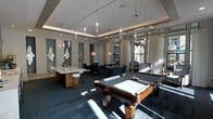 This single-floor amenity lounge is designed to blend recreation and relaxation in a contemporary, well-appointed setting. The space is anchored by a full-size billiards table with rich wood detailing and a slate playing surface, positioned at the center of the room and illuminated by natural light streaming through large windows fitted with sheer white curtains. The tray ceiling adds architectural depth, while an array of pendant lights — ranging from sleek crystal-style drops to warm globe fixtures — creates layered, ambient lighting throughout. A white countertop bar table with modern bar stools provides a casual gathering spot, while a dedicated seating area with navy upholstered chairs and small dining tables offers a more relaxed retreat. Decorative wall-mounted mosaic art panels add visual interest to the neutral grey walls, complementing the space's modern aesthetic. Dark patterned carpet grounds the room, tying together the various zones cohesively. The overall ambiance is polished yet comfortable, making it a functional and attractive space suited for both social interaction and leisure.