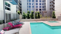 This outdoor aquatic amenity area is situated within a modern urban building complex, surrounded by the striking façades of multi-story buildings that frame the space on multiple sides. The centerpiece is a well-maintained swimming pool with clear turquoise water, complemented by a compact spa or hot tub nearby, both finished with light-toned concrete decking that reflects the sunlight throughout the day.

The pool area is thoughtfully appointed with colorful lounge chairs, offering a relaxed setting for sunbathing and leisure. Tall, manicured trees and lush vertical plantings line the perimeter, softening the urban environment and providing a sense of privacy and greenery within the city landscape. Metal railings and fencing define the boundaries of the space, maintaining a secure and organized layout.

A dedicated shower facility is accessible from the pool deck, conveniently located near a gated entry point, making it easy for guests to transition in and out of the area. Signage throughout the space communicates pool rules and safety information. The overall ambiance balances the energy of a vibrant urban setting with the calm, resort-like feel of a well-designed aquatic retreat.