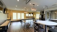 This single-floor community space is designed with both function and style in mind, offering an inviting environment for recreation and socializing. The open layout is anchored by rich hardwood-style flooring and warm yellow accent walls that create an energetic yet welcoming atmosphere. The room features generous ceiling height, enhanced by a combination of recessed lighting and distinctive decorative pendant and chandelier fixtures that add a rustic-industrial character to the space.

The game area is a central highlight, housing a full-size ping-pong table and a pool table, making it an ideal gathering spot for friendly competition. A row of large windows lines the walls, allowing ample natural light to fill the room while providing a bright and airy feel. A comfortable lounge seating area is positioned along the windows, offering a relaxed retreat within the space.

Toward one end of the room, a kitchenette is outfitted with dark cabinetry, a stainless steel refrigerator, and a tiled backsplash, providing convenient amenities for events and gatherings. A round dining table with chairs rounds out the space, adding a casual dining option. Overall, this multi-use recreational room is thoughtfully designed to accommodate a variety of social activities in a comfortable and well-appointed setting.