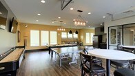 This single-floor community space is designed with both function and style in mind, offering an inviting environment for recreation and socializing. The open layout is anchored by rich hardwood-style flooring and warm yellow accent walls that create an energetic yet welcoming atmosphere. The room features generous ceiling height, enhanced by a combination of recessed lighting and distinctive decorative pendant and chandelier fixtures that add a rustic-industrial character to the space.

The game area is a central highlight, housing a full-size ping-pong table and a pool table, making it an ideal gathering spot for friendly competition. A row of large windows lines the walls, allowing ample natural light to fill the room while providing a bright and airy feel. A comfortable lounge seating area is positioned along the windows, offering a relaxed retreat within the space.

Toward one end of the room, a kitchenette is outfitted with dark cabinetry, a stainless steel refrigerator, and a tiled backsplash, providing convenient amenities for events and gatherings. A round dining table with chairs rounds out the space, adding a casual dining option. Overall, this multi-use recreational room is thoughtfully designed to accommodate a variety of social activities in a comfortable and well-appointed setting.