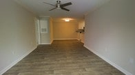 This single-floor space offers a clean, functional layout designed to maximize comfort and usability. The main living area is an open, airy room finished with rich, dark hardwood-style plank flooring that runs throughout, providing a warm and cohesive aesthetic. Crown molding lines the perimeter of the ceiling, adding a refined architectural detail that elevates the overall finish of the space. A ceiling fan with a brushed nickel finish is centrally mounted, contributing both to air circulation and to the room's practical appeal. The walls are painted in a neutral warm tone, creating a bright and welcoming atmosphere. A flush-mount ceiling light fixture in the adjacent area provides additional illumination, guiding the eye toward the kitchen at the far end. The kitchen features white upper cabinetry, dark countertops, and built-in appliances, offering an efficient and organized cooking space. Wide baseboards throughout the unit complement the crown molding, maintaining a consistent and polished interior finish. A hallway leading from the main living area suggests additional rooms beyond, and louvered shutters on a window add a classic touch. Overall, the space presents a well-maintained, move-in-ready environment with thoughtful design details throughout.