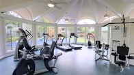 This fitness center occupies a architecturally striking single-story pavilion defined by its signature vaulted, groin-style ceiling — a series of white arched bays that converge overhead to create a dramatic and airy interior. Ceiling fans are mounted at key points beneath the vault, ensuring comfortable air circulation throughout workouts.

The space is lined with large arched windows along its curved perimeter, flooding the interior with abundant natural light and offering unobstructed views of the surrounding green landscape, including a playground area and mature trees. This seamless connection to the outdoors gives the fitness center a bright, open, and inviting atmosphere.

The flooring consists of durable rubber gym matting in a deep charcoal tone, providing both safety and comfort underfoot. The equipment layout is well-organized and includes multiple treadmills, elliptical trainers, a recumbent bike, a seated rowing or cycling machine, and a strength-training cable machine — offering a comprehensive range of cardio and resistance options.

The overall design balances elegant architectural character with practical fitness functionality, making this an exceptional amenity space that stands out for both its form and purpose.