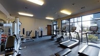 This fitness center offers a comprehensive workout environment designed to support a wide range of exercise routines. The space is thoughtfully laid out across a single level, providing ample room for both cardio and strength training activities. A row of cardio machines — including treadmills, ellipticals, and stationary bikes — lines the perimeter near a large floor-to-ceiling window, allowing natural light to filter in and offering views of an outdoor courtyard area. The free weights section features a full dumbbell rack alongside an adjustable bench, catering to users of varying fitness levels. Additional strength training stations are positioned throughout the room, making efficient use of the available floor space. Durable, dark carpet flooring covers the entire area, providing a practical and comfortable surface underfoot. Overhead, bright fluorescent panel lighting ensures the space is evenly and well-lit at all times. The neutral-toned walls contribute to a clean, focused atmosphere, while a centrally located door provides convenient access to the exterior. A water station is also available, adding to the functionality of the space. Overall, the fitness center is practical, organized, and well-maintained.