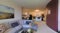 This single-floor residence showcases an open-concept layout that integrates the living, dining, and kitchen areas into one cohesive, flowing space. The living room is anchored by a generously sized sectional sofa in soft gray tones, accented with decorative throw pillows, and complemented by a light-toned carpet and a low-profile coffee table. A piece of wall art adds a creative touch near the entrance.

The dining area sits adjacent to the living space, furnished with a round table and contemporary chairs beneath a striking sputnik-style chandelier that serves as a design focal point. Natural and ambient lighting work together to create a warm, inviting atmosphere throughout.

The kitchen is well-appointed with warm wood-toned cabinetry, a breakfast bar with bar seating, and a full suite of black appliances, including a refrigerator, microwave, and range. A flush-mount ceiling light illuminates the kitchen, while light-colored countertops and a neutral backsplash contribute to the clean, modern aesthetic. The open sightlines between all areas enhance the sense of spaciousness and connectivity throughout the home.