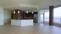 This single-floor space showcases a thoughtfully designed open-plan layout that seamlessly integrates the kitchen and living areas. The kitchen is a standout feature, anchored by a large peninsula with a dark stone countertop and an integrated sink, offering both generous prep space and a natural gathering point. Dark espresso cabinetry lines the walls, complemented by a warm golden-toned brick accent wall that adds texture and character to the space. A stainless steel refrigerator and coordinating appliances reinforce the kitchen's modern aesthetic. Above the peninsula, a series of cylindrical pendant lights hang at varying heights, providing focused task lighting while serving as a decorative element. The flooring throughout is a light-toned large-format tile, lending a clean and cohesive look underfoot. The living area benefits from an abundance of natural light, drawn in through expansive floor-to-ceiling glass doors and windows that frame sweeping panoramic views of the surrounding landscape. This seamless indoor-outdoor connection creates a bright and airy ambiance throughout. The overall design is contemporary and polished, balancing functional kitchen elements with an open, light-filled living environment.
