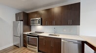This thoughtfully designed single-floor residence offers a clean, contemporary aesthetic throughout. The kitchen is a functional standout, featuring rich dark-wood cabinetry extending to the ceiling for ample storage, white quartz countertops, a light subway tile backsplash, and a full suite of stainless steel appliances including a gas range, microwave, refrigerator, and dishwasher. Recessed lighting and hardwood-style flooring carry through the main living areas, lending warmth and continuity to the open layout. The living area is bright and airy, with a large window framing views of the surrounding streetscape and greenery, and a door providing direct access to an outdoor space. The bedrooms are finished with plush carpet and equipped with generously sized closets outfitted with adjustable wire shelving and hanging rods. The bathroom features a clean, all-white design with large-format marble-look floor tiles, a soaking tub with tiled surround, and a vanity with dark wood cabinetry and a white countertop. A dedicated in-unit laundry closet houses a full-size stacked washer and dryer. The overall ambiance is modern and move-in ready, combining practical amenities with stylish finishes throughout.