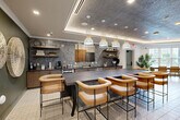 Located at 515 Royal Court in uptown Charlotte, NC, this thoughtfully designed amenity lounge offers a generously sized, single-floor layout that seamlessly blends social, dining, and entertainment functions. The space is anchored by a striking bar and kitchen area, finished with textured gray stone tile backsplash, warm wood cabinetry, open shelving with curated décor, and a row of elegant pendant lights suspended above a substantial dark-topped counter. Cognac leather bar stools with wood frames provide comfortable seating throughout.

The lounge transitions into a bright seating corridor lined with large windows that invite abundant natural light and views of the surrounding Charlotte skyline. Tall tropical plants and patterned tile flooring contribute to the sophisticated, resort-inspired ambiance. A bold botanical-print feature wall anchors a lounge zone furnished with a sectional sofa and additional bar-height tables, while woven wall art and a statement chandelier add texture and warmth to adjacent dining areas.

A dramatic tray ceiling with integrated lighting, textured metallic ceiling panels, and architectural columns further define the space. Rounding out the offerings is a dedicated Full Swing golf simulator room, providing a premium entertainment experience within this well-appointed amenity space.