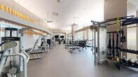 Located at 575 12th Road South in Arlington, VA, this fitness center offers a generous, thoughtfully designed space across two dedicated exercise rooms. The facility features a sophisticated architectural design, highlighted by soaring ceilings, a striking curved wood-slat ceiling detail in the studio area, and large floor-to-ceiling windows that flood the space with natural light and provide views of the surrounding neighborhood.

The main exercise room is outfitted with an extensive selection of strength and cardio equipment, including cable machines, multi-use weight stations, ellipticals, stationary bikes, treadmills, and a full rack of dumbbells. Durable rubber flooring throughout provides comfort and safety during workouts, while wall-to-wall mirrors enhance the sense of space and allow for proper form checks. A decorative textured feature wall adds a refined visual element to the room.

The second exercise room serves as a versatile studio space with warm hardwood flooring, full-length mirrors, spinning bikes, rowing machines, medicine balls, and a wall-mounted flat-screen TV — ideal for group classes or individual training sessions. A ceiling fan and recessed lighting complete the bright, airy ambiance. Together, both rooms deliver a comprehensive and modern fitness experience in an upscale, well-appointed setting.