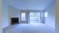 This single-floor residence offers a welcoming and well-lit living space designed for comfort and functionality. The main living area is finished with soft, plush wall-to-wall carpeting that adds warmth underfoot and a sense of coziness throughout the room. A fireplace with a built-in media console serves as a natural focal point, providing both a practical and aesthetic centerpiece to the space.

Natural light plays a significant role in the ambiance of this home. Multiple windows along the exterior walls allow daylight to filter in from multiple directions, keeping the interior bright and airy. Neutral gray tones on the walls complement the light carpeting, creating a clean and versatile palette suited to a variety of interior styles.

A set of sliding glass doors opens directly onto a private balcony, offering a seamless transition between indoor and outdoor living. The balcony features a white railing and is surrounded by lush greenery, providing a serene and natural backdrop. The combination of abundant windows, the sliding door, and the outdoor views gives the living area an open, expansive feel. Overall, the space presents a calm and inviting atmosphere ideal for everyday living.