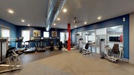 This fitness center is a thoughtfully designed workout facility that combines functionality with a modern aesthetic. The space is defined by bold navy blue walls that create an energized atmosphere, complemented by a striking red structural column that adds a pop of industrial character. Exposed silver ductwork runs along the ceiling, contributing to the space's urban, warehouse-inspired design, while recessed lighting and a ceiling fan ensure the environment remains well-lit and comfortable during workouts.

The main workout floor is outfitted with a variety of cardio machines, including treadmills and ellipticals equipped with individual digital screens for an interactive fitness experience. Strength training is well-represented with cable and weight machines positioned throughout the floor, offering users a comprehensive full-body workout option. The flooring transitions between durable rubberized material and a neutral-toned surface, providing both safety and comfort underfoot.

Natural light filters in through windows along one wall, while a glass-paneled partition wall on the opposite side adds an open, airy quality to the space. Motivational artwork displayed on the walls rounds out the environment, making this fitness center a practical and inviting destination for all levels of fitness enthusiasts.