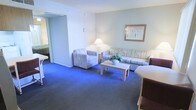 This single-floor suite offers a well-appointed and functional layout designed for comfort and relaxation. The living area serves as the heart of the space, featuring plush, patterned upholstered seating including a sofa and an armchair, arranged around a light wood coffee table — ideal for unwinding or casual conversation. Soft blue wall-to-wall carpeting runs throughout, adding a calming, cohesive feel to the interior. Neutral light-grey walls keep the atmosphere bright and airy, complemented by a wall-mounted lamp and a classic table lamp that provide warm, layered lighting. A framed piece of wall art adds a decorative touch to the room. An open corridor visible in the background connects to additional areas of the suite, suggesting a thoughtfully separated sleeping or bathroom space beyond. The overall design reflects a clean, classic hospitality aesthetic, balancing practicality with comfort. The space is well-suited for extended stays or relaxed accommodations, offering a living environment that feels both organized and inviting.