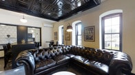 Located at 345 East Deshler Avenue in Columbus, Ohio, this single-floor space delivers a distinctly refined and characterful interior design. The main lounge area immediately captures attention with its dramatic coffered tin ceiling finished in a deep black, lending the room a bold, vintage aesthetic. A generously sized tufted leather sectional sofa anchors the seating area, offering a classic Chesterfield-style elegance that complements the overall design language. Arched windows with white trim frame natural light beautifully, contrasting the darker ceiling and furniture tones and creating a well-balanced ambiance. The warm, cream-toned walls serve as a neutral backdrop, allowing the darker architectural and furnishing elements to stand out. Framed vintage-style artwork adorns the walls, adding a layer of cultural and historical character to the space. A wall-mounted split air conditioning unit ensures modern climate comfort without disrupting the classic aesthetic. An adjacent area, partially visible through a decorative partition, features additional seating and dining arrangements, suggesting a versatile layout suited for both relaxed gatherings and more structured events. The overall atmosphere is sophisticated, warm, and rich in detail.