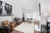 This thoughtfully designed two-floor residence combines modern finishes with a warm, inviting ambiance throughout its well-proportioned layout. The main living area is anchored by soaring vaulted ceilings and rich hardwood flooring that extends seamlessly through the space, creating an open and airy atmosphere. A striking fireplace serves as a focal point in the living room, which flows toward a bay window framing lush, mature greenery outside. The kitchen features white cabinetry, stainless steel appliances, a built-in breakfast bar with counter seating, and recessed lighting overhead. A versatile sitting or study area offers additional functional space, complete with vaulted ceilings and a statement pendant light. Multiple bathrooms are finished with clean white vanities, large mirrors, and wood-look flooring, maintaining a consistent contemporary aesthetic. The spacious bedroom is carpeted for comfort and benefits from generous natural light streaming through wide windows that overlook verdant trees, providing a serene retreat. Throughout the residence, a neutral color palette of whites and warm tones ties each area together, while curated wall art and thoughtful décor add character and visual interest at every turn.