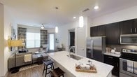 This single-floor residence showcases a thoughtfully designed open-concept layout that integrates the kitchen and living areas into one cohesive, flowing space. The kitchen serves as a central highlight, featuring a large granite-topped island with an undermount sink and seating for multiple guests — ideal for both meal preparation and casual dining. Dark cabinetry contrasts elegantly with the light stone countertops, while stainless steel appliances, including a French door refrigerator with ice dispenser, add a contemporary, polished finish. Pendant lighting with cylindrical white shades hangs above the island, providing focused illumination and a modern aesthetic touch.

The living area, visible beyond the kitchen, benefits from abundant natural light streaming through large windows dressed with patterned curtains. A ceiling fan with integrated lighting ensures comfortable airflow throughout. Wood-look flooring runs continuously across both spaces, reinforcing the open and unified design. Neutral walls and recessed lighting further enhance the bright, airy ambiance. A doorway visible in the background suggests additional private rooms, rounding out this well-appointed and functional single-level residence.
