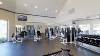 Located in Arlington, Texas, this fitness center offers a comprehensive workout environment designed to accommodate a wide range of exercise needs. The facility is housed on a single floor beneath soaring vaulted ceilings that lend an open, airy feel to the entire space. Recessed lighting illuminates the room evenly, ensuring a bright and energizing atmosphere throughout.

The main workout area is outfitted with an extensive selection of strength training machines, including cable systems, press stations, and resistance equipment, primarily finished in a sleek black-and-white color scheme. Cardio machines are also available, positioned to provide a well-rounded fitness experience. Full-length mirrors line the walls, amplifying the sense of space while allowing members to monitor their form during exercise.

Durable dark flooring runs throughout the facility, providing a practical and professional surface suited for high-traffic use. Mounted flat-screen televisions are positioned at multiple points within the room, offering entertainment or instructional content during workouts. Large windows allow natural light to filter into the space, complementing the recessed overhead lighting. The overall ambiance is clean, modern, and purposefully designed to support an effective and comfortable fitness experience.