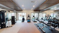 Located at 1602 Belt Line Rd in Garland, TX, this modern fitness facility is thoughtfully designed to support a comprehensive workout experience across a single, well-organized floor. The exercise room serves as the centerpiece of the space, outfitted with a wide array of professional-grade equipment including TRUE-brand treadmills and ellipticals, multi-station cable machines, free weight racks with dumbbells, adjustable benches, and functional training accessories. A distinctive wood-slat ceiling detail anchors the room's contemporary aesthetic, while floor-to-ceiling mirrors visually expand the space and allow users to monitor their form throughout their workouts. Recessed lighting provides consistent illumination throughout. Adjacent to the main gym floor is a dedicated movement studio with warm wood-look flooring, large mirrored walls, and medicine ball storage — ideal for stretching, yoga, or group exercise. Large sliding glass doors and expansive windows throughout the facility bring in generous natural light and offer views of the outdoor surroundings, including what appears to be a pool area. The overall ambiance is clean, bright, and modern, creating a motivating environment for residents and users seeking a full-featured fitness experience.
