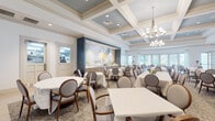 Located at 3650 RCA Blvd in Palm Beach Gardens, Florida, this single-floor community clubhouse offers a well-appointed collection of social and recreational amenities designed for comfort and elegance. The formal dining room is a standout feature, showcasing coffered ceilings accented in soft blue and white with decorative crown molding, elegant chandelier lighting, and a large-scale floral mural that anchors the space. Neatly dressed tables with white linens and rounded upholstered chairs fill the room, while abundant natural light filters through multiple windows. A buffet service counter is integrated seamlessly into the dining area. Adjacent to the dining room, the lounge and bar area features a sweeping curved bar with a quartz countertop, blue mosaic tile detailing, built-in shelving, and stylish pendant lighting. This zone also includes a billiards table and a wall-mounted television, creating a lively social hub. French doors open to the outdoors, enhancing the airy atmosphere. A dedicated game room with a round poker table sits within a curved bay of floor-to-ceiling windows, bathing the space in natural Florida sunlight and offering views of the surrounding landscape. Light wood-tone flooring flows throughout, unifying the entire facility with a clean, modern coastal aesthetic.