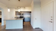 This single-floor, one-bedroom, one-bathroom residence offers a thoughtfully designed layout that balances functionality and modern style. The kitchen serves as a standout feature, equipped with stainless steel appliances including a gas range, built-in microwave, and full-size refrigerator. Dark-toned cabinetry with brushed metal hardware pairs with granite countertops and a white subway tile backsplash to create a contemporary aesthetic. A peninsula with matching granite provides additional prep space and casual seating potential. Track lighting and a pendant fixture illuminate the kitchen, complementing the warm wood-look hardwood flooring that extends through the main living areas. Crown molding adds an elevated architectural detail to the space. The bedroom features plush neutral carpet, creating a comfortable retreat, and includes a window that allows natural light and outdoor views. The bathroom continues the design consistency with dark cabinetry, granite countertops, wood-look flooring, and a full tub-shower combination. The 3D overview reveals an open-concept connection between the kitchen and living area, with high ceilings and sliding glass door access that enhances the sense of spaciousness throughout.