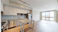 This contemporary one-bedroom, one-bathroom residence at 880 Pacific Street in Stamford, Connecticut offers a thoughtfully designed open-concept layout with modern finishes throughout. Warm wood-tone flooring flows seamlessly across the entire unit, complementing the neutral color palette and creating a cohesive, welcoming ambiance.

The kitchen and living area share an open, connected space ideal for everyday living and entertaining. The kitchen is outfitted with light wood cabinetry extending to the ceiling, a soft blue-gray tile backsplash, stainless steel appliances including a gas range, built-in microwave, dishwasher, and French door refrigerator, as well as a central island that doubles as a dining surface. Track lighting above adds a modern, functional touch.

The living area is bright and airy, anchored by a large floor-to-ceiling sliding glass window that frames views of the surrounding Stamford skyline and neighboring green spaces below, filling the room with natural light.

The spacious bedroom continues the light, open feel with its own generous floor-to-ceiling window offering additional city and park views. Two closets provide ample storage throughout the unit. The full bathroom features a soaking tub with tiled surround, a contemporary vanity with an illuminated mirror, and wood-look tile flooring. An in-unit laundry area completes this well-appointed residence.