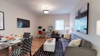This single-floor residence offers a thoughtfully designed layout that balances comfort and functionality. The open-concept living and dining area is bright and welcoming, anchored by warm hardwood flooring and filled with natural light from a large window fitted with vertical blinds. The living space accommodates a tufted gray sofa, armchairs, and a white coffee table set atop a bold black-and-white polka dot rug, while a wall-mounted flat-screen TV and a dedicated desk area with shelving add both entertainment and work-from-home convenience. The dining area flows seamlessly from the living space, featuring a stylish table and patterned chairs. The kitchen is well-equipped with light maple cabinetry, granite-style countertops, and a full suite of stainless steel appliances including a refrigerator, range, microwave, and dishwasher, all set on neutral tile flooring. The bedroom is a serene retreat with plush carpeting, a spacious closet, and room for a full dresser and bedside storage. The bathroom features a granite-topped vanity with wood cabinetry, an oval mirror, and clean, neutral finishes throughout. The overall ambiance is modern, clean, and move-in ready.