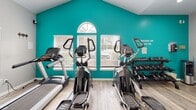 Located at 1200 Virginia Center Pkwy in Glen Allen, Virginia, this fitness center offers a well-equipped, single-floor workout facility designed to support a full range of exercise routines. The space is accessed through a glass-paneled entry door flanked by white plantation shutters, creating a welcoming and polished first impression.

Inside, the gym is defined by vaulted ceilings that rise to a striking arched window, flooding the main workout area with natural light. Bold teal accent walls contrast with neutral gray and white surfaces, lending the space an energetic and motivating atmosphere. Light wood-style flooring runs throughout, providing a clean and durable foundation underfoot.

The cardio section is well-appointed with multiple treadmills and elliptical machines positioned to take advantage of the natural light from the windows. Strength training needs are addressed with a multi-function cable machine, a dumbbell rack stocked with a range of free weights, and a collection of medicine balls on a dedicated storage rack. A yoga mat is also available for stretching and floor exercises.

Recessed lighting supplements the natural light in areas with lower ceiling heights, ensuring the entire space remains bright and functional. Motivational signage adorns the walls, reinforcing the fitness-focused atmosphere. The layout is thoughtfully organized to provide clear circulation between equipment zones, making this facility practical and accessible for all fitness levels.