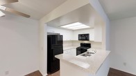 This single-floor space is designed with functionality and style in mind. The kitchen serves as a central highlight, featuring crisp white cabinetry that extends to the ceiling and provides ample storage. A peninsula with light granite countertops offers additional workspace and creates a natural division between the kitchen and the adjoining area, making it ideal for casual dining or entertaining. The kitchen is equipped with a full suite of black appliances, including a refrigerator, gas range, over-the-range microwave, and a stainless steel sink. A recessed skylight panel overhead floods the cooking area with natural light, enhancing the bright, airy atmosphere created by the white walls and cabinetry. The clean lines and neutral color palette throughout give the space a timeless, modern feel. Hardwood-style flooring adds warmth and continuity, grounding the overall aesthetic. The open sightlines and thoughtful layout make the space feel welcoming and practical, catering to everyday living with ease.