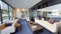 This elegantly appointed single-floor space showcases a masterfully designed contemporary interior that exudes luxury and refinement. The main living area is anchored by a plush white sofa adorned with navy and ivory accent pillows, complemented by a pair of matching white armchairs that create an inviting conversational arrangement. A substantial low-profile coffee table in a dark finish serves as the room's centerpiece, styled with decorative bronze bowls and coffee table books. The space features rich blue-grey carpeting that adds warmth and depth underfoot, while recessed lighting throughout provides a clean, modern ambiance. A striking dark wood and lacquer feature wall with integrated cabinetry and shelving adds dramatic contrast to the lighter furnishings. Full-height mirrored panels expand the sense of space and reflect light throughout the room, enhancing its open and airy feel. Slender black side tables with metallic accents add functional elegance alongside the seating. Sheer curtains allow soft, diffused natural light to filter in, contributing to the space's calm and sophisticated atmosphere. The overall design aesthetic is polished and cohesive, blending dark architectural elements with light furnishings for a balanced, high-end look.
