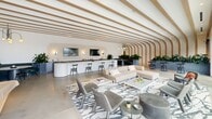 Located at 320 W South St in downtown Raleigh, NC, this thoughtfully designed amenity space spans a generous single floor, offering a refined collection of social, wellness, and recreational areas. The centerpiece is a sophisticated lounge defined by striking arched wood ceiling ribs that sweep across the expanse, polished concrete floors, and abundant natural light streaming through floor-to-ceiling windows. A curved white bar with counter seating anchors one side, complemented by plush sectional sofas, accent chairs, and bold patterned area rugs. Lush indoor planters add vibrant greenery throughout, creating a welcoming, resort-like atmosphere. A well-appointed corridor connects seamlessly to the fitness center, which is divided into distinct zones featuring strength machines, cable rigs, boxing equipment, cycling bikes, treadmills, TRX systems, and floor-to-ceiling mirrors — all beneath bright linear lighting with panoramic city views. The outdoor areas elevate the experience further with a resort-style rooftop pool flanked by cabana loungers and open sky views of the Raleigh skyline and surrounding landscape. An outdoor kitchen and entertaining terrace, covered by a sleek pergola, features a chevron-tiled accent wall, built-in grills, a bar counter, and a mounted TV — ideal for gatherings in an elevated urban setting.