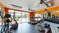 Located at 50 Maner Ter SE in Smyrna, Georgia, this fitness center is a well-equipped, single-room exercise facility designed to serve the needs of active residents. The space is defined by its bold orange accent walls, which lend an energetic and motivating atmosphere, complemented by durable dark rubber flooring suitable for high-traffic workout use.

The room is furnished with a comprehensive selection of exercise equipment, including multiple treadmills, an elliptical trainer, a recumbent bike, an upright stationary bike, and a multi-station cable weight machine, along with a standalone pull-up and dip tower. A full-length wall mirror runs along one side of the room, visually expanding the space and allowing users to monitor their form during workouts.

Natural light floods the fitness center through expansive floor-to-ceiling sliding glass windows and doors along one wall, offering direct views of the outdoor pool area and surrounding landscaped grounds. This connection to the outdoors enhances the overall ambiance and keeps the space feeling open and airy. Ceiling fans are mounted overhead to support airflow and comfort during exercise sessions. The facility provides a focused, practical environment with everything needed for a complete cardio and strength training workout.