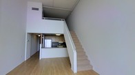 This two-floor loft-style space showcases an industrial-modern design defined by soaring double-height ceilings with exposed structural elements overhead. The main level features light hardwood flooring that runs throughout, lending warmth to the otherwise crisp, neutral palette of white and grey tones. A compact, well-appointed kitchen sits neatly tucked into the main living area, equipped with light wood cabinetry, dark countertops, a full-size refrigerator, and under-cabinet task lighting. The open-plan layout of the main floor creates a generous sense of space and flow.

A centrally positioned staircase with a clean white railing leads to the upper level, where an open loft overlooks the main living area below through a metal railing. This mezzanine level benefits from the dramatic vertical volume created by the high ceilings and exposed ceiling infrastructure, giving the space an airy, urban character.

A bright corridor lined with white walls and light hardwood floors connects the various areas of the unit, illuminated by a flush-mount ceiling fixture. Natural light filters in at the end of the hallway, contributing to the overall bright, open ambiance of this thoughtfully designed multi-level space.