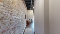 This 1-bedroom, 1-bathroom loft-style unit in Oxford, MI, blends industrial character with modern finishes throughout its thoughtfully designed single floor. Soaring ceilings with exposed beams, steel ductwork, and dark-painted structural elements define the open living area, while expansive original brick walls add warmth and texture throughout the space. Rich hardwood flooring flows seamlessly from the entry corridor through the main living and sleeping areas.

The kitchen is a standout feature, centered around a large island with bar seating for four, complemented by gray shaker cabinetry, white subway tile backsplash, stainless steel appliances, and bold brass pendant lighting that adds a refined industrial touch. The open-concept layout connects the kitchen directly to the living and sleeping areas, maximizing the sense of space.

The bathroom suite is well-appointed, featuring gray vanity cabinetry with gold hardware, a walk-in glass shower with white subway tile walls, and a classic black-and-white hexagonal mosaic tile floor. A separate water closet provides added privacy. The dedicated laundry room includes a full-size front-loading washer and dryer with overhead gray cabinetry for additional storage. Large windows throughout invite natural light and offer views of the surrounding Oxford streetscape.