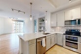 Located in the vibrant South End neighborhood of Charlotte, NC, this 2-bedroom, 2-bathroom residence offers a thoughtfully designed single-floor layout with contemporary finishes throughout. The open-concept kitchen and living area serves as the heart of the home, featuring gray shaker cabinetry, quartz countertops, a herringbone tile backsplash, stainless steel appliances, and a peninsula island with pendant lighting — all flowing seamlessly into the living space with warm hardwood flooring. Built-in open shelving adds a stylish, functional accent. The living area is bathed in natural light from expansive floor-to-ceiling windows that frame elevated views of the surrounding Charlotte skyline. The primary bedroom offers plush carpeting, a ceiling fan, and direct access to a private balcony with city views, while the second bedroom features hardwood floors and a distinctive clerestory window detail. The primary bathroom is finished with large-format travertine tile, a quartz vanity, framed mirror, glass-enclosed shower, and access to a walk-in closet. The second bathroom echoes the same refined palette with a white quartz vanity and gold-accented mirror. A connecting hallway ties the layout together efficiently, completing this polished urban residence.