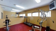 This fitness room is a dedicated exercise space designed to support a well-rounded workout routine. The room features warm yellow walls, a bold red carpet underfoot, and bright fluorescent lighting that ensures the space is well-illuminated at all times. A row of windows along one wall, fitted with white horizontal blinds, allows for natural light while maintaining privacy.

The space is equipped with a treadmill for cardiovascular exercise and a multi-station strength training machine, providing users with options for both aerobic and resistance-based workouts. A wall-mounted television is positioned for convenient viewing during exercise sessions. The mirrored wall along one side of the room visually expands the space and allows users to monitor their form during workouts.

The single-floor layout is straightforward and functional, maximizing the available footprint for equipment use. The combination of practical flooring, reflective surfaces, and adequate natural and artificial lighting creates a utilitarian yet comfortable environment suited for regular fitness activity.