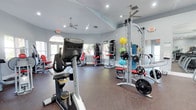 This athletic club offers a comprehensive fitness environment designed to support a wide range of workout routines. The space features vaulted ceilings that contribute to an open, airy atmosphere, complemented by ceiling fans that promote air circulation throughout. Recessed lighting, combined with natural light streaming through arched windows, ensures the facility is well-lit and welcoming.

The workout floor is outfitted with commercial-grade Matrix brand equipment, including a stationary bike and additional cardio machines positioned toward the front of the space. A multi-station cable and weight machine system anchors the strength-training area, accompanied by plate-loaded equipment and adjustable benches. Colorful stability and medicine balls are also available for functional training exercises.

Rubber flooring covers the workout area, providing durability and impact absorption suitable for a high-use fitness environment. Full-length mirrors line one wall, allowing users to monitor their form during exercise. The walls are finished in a neutral gray tone, giving the space a clean, modern aesthetic. Emergency exit signage is clearly visible, reflecting attention to safety throughout the facility.