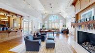 Located at 1000 Bally Bunion Dr in Egg Harbor City, NJ, this well-appointed single-floor community space combines architectural elegance with comfortable, functional design. The centerpiece of the main gathering area is its dramatic vaulted ceiling, which rises to impressive heights and is complemented by a striking wall of arched, multi-pane windows that flood the interior with abundant natural light and frame lush outdoor views of the surrounding neighborhood. Warm hardwood flooring runs throughout the open main area, anchored by a handsome wood-trimmed fireplace that serves as a natural focal point for relaxed seating arrangements featuring blue upholstered sofas and chairs. Track lighting along the ceiling and pendant fixtures add warmth during evening hours. French doors provide seamless access to the outdoors, enhancing the bright and airy ambiance. A dedicated kitchen area is conveniently accessible via a crafted wood-detailed service counter. The space also includes a game room, outfitted with a billiards table, high-top seating, striped carpeting, and wood-framed artwork, offering a more casual and recreational retreat. Together, these spaces create an inviting environment well-suited for both social gatherings and leisurely activities.