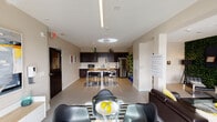 This single-floor space is designed with an open-concept layout that seamlessly connects the kitchen, dining, and lounge areas, creating a cohesive and functional environment. The kitchen is outfitted with dark espresso cabinetry, white countertops, stainless steel appliances including a full-size refrigerator, microwave, and dishwasher, along with ample upper and lower storage. Recessed lighting illuminates the kitchen and dining zone, where a wood-topped dining table with modern metal-framed stools provides a practical gathering spot.

The lounge area is styled with a dark leather sofa accented by yellow throw pillows, a glass-top round dining table surrounded by contemporary black chairs, and a bold black-and-white striped area rug. A large sliding glass door floods the space with natural light and opens to an outdoor terrace with urban views. Decorative touches include a striking large-scale abstract artwork, a living green wall feature, and indoor potted plants that add organic warmth.

Linear pendant lighting runs across the ceiling, complementing the neutral warm-gray walls and light tile flooring. The overall ambiance is modern and stylish, balancing bold design accents with a clean, functional aesthetic suited for communal or multi-purpose use.