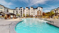 Located at 10051 Perimeter Pkwy in Charlotte, NC, this outdoor amenity space offers a resort-inspired experience centered around a generously sized swimming pool with striking turquoise water. The pool features a freeform design with multiple entry points via stainless steel handrails and a defined 5-foot depth zone, accommodating both casual swimmers and those seeking relaxation.

Surrounding the pool is an expansive stamped stone paver deck that provides ample room for lounging and socializing. The deck is outfitted with rows of modern white-framed chaise lounges with gray slatted surfaces, paired with coordinating side tables and shaded umbrella stations, creating comfortable outdoor retreats throughout the space. Lush landscaping with ornamental grasses, evergreen shrubs, and decorative rock beds adds natural texture and greenery to the environment.

A standout architectural feature is the covered outdoor pavilion, constructed with a pitched roof, timber framing, and stone accents. This structure provides shaded seating and serves as a central gathering point within the courtyard. The entire amenity area is enclosed within a multi-story residential community, with brick and light-toned building facades rising around the space, offering a sense of privacy while contributing to the vibrant, community-oriented atmosphere of this Charlotte property.