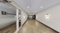 This single-floor lobby space serves as the main entry and circulation hub of a modern residential building. The layout encompasses two distinct zones: a welcoming entrance hall and an inner corridor leading to key building amenities.

The entrance hall greets visitors with large floor-to-ceiling glass doors and windows, flooding the space with natural light and offering clear sightlines to the exterior. Light grey large-format floor tiles, recessed ceiling lighting, and crisp white walls adorned with classic raised panel molding create a clean, sophisticated aesthetic throughout.

Moving deeper into the space, the corridor transitions to warm-toned stone-look floor tiles, maintaining visual continuity while adding depth. This area houses a pair of elevator banks framed by dark grey marble-style wall cladding, providing a striking contrast to the bright white paneled walls. Wall-mounted sconces offer warm accent lighting along the corridor.

Adjacent to the elevators, a dedicated mailbox and parcel station provides residents with convenient package and mail access. A glazed door leads to what appears to be a leasing or management office, identifiable by its signage and interior seating visible through the glass. The overall ambiance is polished and functional, balancing residential comfort with a professionally finished, well-maintained environment.