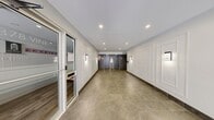 This single-floor lobby space serves as the main entry and circulation hub of a modern residential building. The layout encompasses two distinct zones: a welcoming entrance hall and an inner corridor leading to key building amenities.

The entrance hall greets visitors with large floor-to-ceiling glass doors and windows, flooding the space with natural light and offering clear sightlines to the exterior. Light grey large-format floor tiles, recessed ceiling lighting, and crisp white walls adorned with classic raised panel molding create a clean, sophisticated aesthetic throughout.

Moving deeper into the space, the corridor transitions to warm-toned stone-look floor tiles, maintaining visual continuity while adding depth. This area houses a pair of elevator banks framed by dark grey marble-style wall cladding, providing a striking contrast to the bright white paneled walls. Wall-mounted sconces offer warm accent lighting along the corridor.

Adjacent to the elevators, a dedicated mailbox and parcel station provides residents with convenient package and mail access. A glazed door leads to what appears to be a leasing or management office, identifiable by its signage and interior seating visible through the glass. The overall ambiance is polished and functional, balancing residential comfort with a professionally finished, well-maintained environment.