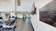 This fitness center offers a thoughtfully designed workout environment within a single open floor plan. The space is defined by soaring double-height ceilings and expansive floor-to-ceiling windows that flood the interior with abundant natural light and provide views of the surrounding outdoor area, creating an energizing atmosphere for exercise.

The facility is comprehensively outfitted with a wide range of equipment to support diverse fitness routines. The cardio zone features multiple treadmills, elliptical trainers, stationary bikes, and a rowing machine, all arranged to maximize usable floor space. The strength training area includes a multi-station cable machine, weight benches, and additional resistance equipment. Full-length mirrors line portions of the walls, enhancing the sense of openness and allowing users to monitor their form during workouts.

Durable, dark-toned flooring provides a practical and resilient surface throughout. Recessed lighting supplements the natural light from the windows, ensuring the space is well-lit at all times. A wall-mounted flat-screen television is positioned for easy viewing from the cardio equipment. Posted fitness guidelines and instructional materials are visible along the walls. A hallway with additional utility space, including storage and restroom access, is accessible from the main workout area, adding to the overall functionality of this well-appointed fitness facility.