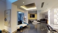 This single-floor space showcases a thoughtfully designed contemporary interior that blends clean lines with bold architectural details. The polished concrete flooring runs throughout, reflecting light and reinforcing the modern aesthetic while providing a durable, low-maintenance surface. A prominent coffered ceiling with warm recessed lighting serves as a focal point, adding depth and a refined sense of structure to the main living area.

The open-plan layout seamlessly connects the lounge area to adjacent spaces, including what appears to be a kitchenette or wet bar visible in the background. The lounge is anchored by a wall-mounted flat-screen television set against a yellow accent wall with geometric hexagonal paneling, flanked by sleek gray upholstered seating and a glass-top coffee table arranged on a patterned area rug.

A floor-to-ceiling sliding glass door floods the interior with natural light and provides direct access to an outdoor balcony or terrace, enhancing the sense of openness. A tufted bench and a metallic accent stool near the entrance add both function and style. The overall ambiance is sophisticated, modern, and well-appointed.