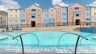 This residential amenity space offers a comprehensive fitness and recreation experience designed to support an active lifestyle. The fitness center is a standout feature, outfitted with professional-grade Life Fitness equipment throughout multiple dedicated zones. One area houses a row of cardio machines, including treadmills and rowing machines, positioned beneath large windows that fill the space with natural light. A separate strength training zone features a robust multi-station cable and functional training rig, a full dumbbell rack, medicine balls, and TRX suspension equipment — providing a thorough free weights and resistance training environment. The rubber flooring, recessed linear lighting, and blue accent walls lend the gym a modern, clean aesthetic. Additional amenities include wall-mounted yoga mat storage and televisions for entertainment during workouts. Entry to the fitness center is through a set of glass double doors, offering visibility into the bright interior. Outside, a generously sized outdoor swimming pool with crystal-clear water serves as the centerpiece of the recreational area. The pool is framed by a paved deck, iron railings, and a striking multi-story residential building clad in alternating panels of coral, salmon, and blue siding — creating a vibrant and welcoming community atmosphere under open skies.