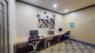 This business center is a thoughtfully designed workspace that efficiently accommodates multiple users within a compact footprint. A continuous stone-topped counter runs along the main wall, providing ample desk surface for two computer workstations equipped with desktop monitors and ergonomic task chairs. A shared printer sits centrally on the counter, offering convenient access to all users. A private nook at one end of the room features an additional workstation with its own monitor and chair, ideal for users seeking a quieter work environment.

The room is finished with neutral warm-beige walls accented by dark crown molding and a coordinating baseboard detail, lending the space a polished, professional aesthetic. Recessed ceiling lighting provides consistent, even illumination throughout. The floor is covered in a bold patterned carpet featuring gold swirl motifs on a dark background, adding visual interest and a contemporary touch.

Decorative wall art depicting close-up typewriter key imagery, along with sculptural starburst wall accents, contribute a creative, design-forward character to the room. A set of glass-paneled double doors connects the space to an adjacent fitness area, enhancing the sense of openness. Posted usage guidelines are clearly displayed, reinforcing the room's functional, shared-use purpose.