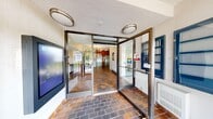 This single-floor lobby and entrance area offers a well-organized and functional layout spanning a generously sized footprint. The space is accessed through three distinct entrance points, each framed by full-height glass doors and metal framing that allow abundant natural light to flow inward, providing clear sightlines to the surrounding outdoor greenery.

The interior is anchored by warm-toned brick-patterned tile flooring that extends throughout the main lobby and connecting hallway, lending a classic, durable character to the space. An elegant multi-arm chandelier serves as a focal point overhead, complementing the elevated ceiling height in the central lobby area and adding a refined aesthetic contrast to the textured ceiling finish.

Practical amenities are thoughtfully integrated throughout. A stainless steel elevator provides vertical access, while a built-in mailbox and parcel station serves residents or occupants efficiently. A seating area with leather armchairs and a console table offers a welcoming rest point. A dedicated small office or workstation room, finished with light tile flooring and recessed lighting, provides a quiet workspace complete with a desk, seating, and printer. Flush-mount ceiling fixtures and baseboard heating units complete the functional infrastructure of this well-appointed lobby space.