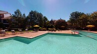 Located at 2500 Grove Club Ln in Cary, NC, this single-level outdoor amenity space is thoughtfully designed to offer a resort-like experience. At its centerpiece is a generously sized swimming pool with crystal-clear water, framed by a wide concrete pool deck furnished with lounge chairs and colorful umbrellas. Towering pine trees and lush landscaping provide a natural backdrop, creating a serene and private atmosphere under open blue skies.

Adjacent to the pool is an expansive composite deck that extends the outdoor living area, offering multiple seating arrangements including shaded dining sets beneath bright yellow umbrellas. The deck connects seamlessly to a covered pavilion, which serves as a standout architectural feature. The pavilion boasts a vaulted wood-paneled ceiling, lattice railing details, and a striking floor-to-ceiling stone fireplace with a mounted television above — ideal for year-round outdoor enjoyment. The brick exterior building nearby complements the warm, traditional aesthetic of the space.

Well-maintained gardens, ornamental shrubs, and blooming crape myrtles add color and visual interest throughout, while the overall layout encourages both relaxation and social gatherings. This amenity space delivers a polished, community-focused outdoor environment with notable architectural character.