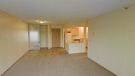 This single-floor residence offers a clean, functional layout designed for comfortable everyday living. The spacious living area is finished with plush wall-to-wall carpeting, creating a warm and inviting atmosphere that anchors the main gathering space. A pass-through kitchen opens directly to the living area, promoting an open-concept feel. The kitchen is equipped with white cabinetry, a refrigerator, a dishwasher, and ample counter space, all illuminated by warm overhead lighting. The transition between the kitchen and living area is defined by a raised breakfast bar-style partition, adding both visual separation and utility. Near the front entry, tile flooring provides a practical and durable surface. A wood-toned entry door welcomes residents and guests, flanked by a generously sized closet that offers convenient storage. Adjacent to the entry, a mirrored closet further enhances the sense of space while providing additional storage. Neutral wall tones throughout lend a versatile backdrop that suits a variety of décor styles. The overall ambiance is bright, understated, and move-in ready, making this a practical and welcoming single-floor living space.