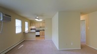 This single-floor space presents an open-concept layout connecting the living area to the kitchen, creating a functional and cohesive environment. The living area is finished with neutral-toned carpet, contributing to a warm and comfortable atmosphere, while the walls are painted in a soft, light yellow-beige tone that enhances the brightness of the space.

The kitchen is partially separated by a half-wall peninsula, which doubles as a breakfast bar or prep surface. It features light wood cabinetry providing ample storage, dark countertops, and white appliances including a gas range and refrigerator. The kitchen floor transitions to hardwood-style flooring, offering a practical surface in the cooking area. A window positioned above the sink allows natural light to filter into the kitchen.

A ceiling fan is centrally mounted overhead, providing both lighting and air circulation across the open space. Baseboard heating units are visible along the walls, offering an efficient heating solution. The overall design is straightforward and functional, with clean lines and a neutral palette that offers versatility for various furnishing styles.