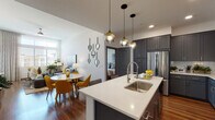 This well-appointed single-floor residence combines modern design with functional living across a thoughtfully arranged open floor plan. The kitchen serves as a central highlight, featuring floor-to-ceiling dark gray shaker-style cabinetry, crisp white quartz countertops, a subway tile backsplash, and a full suite of stainless steel appliances. A generously sized center island with a dual-basin sink and pendant globe lighting adds both workspace and a natural gathering point that flows directly into the dining area. Rich hardwood flooring extends throughout the main living areas, connecting the kitchen and dining space to the living room, which benefits from oversized windows that draw in abundant natural light and elevate the sense of height and openness. The living room offers a welcoming, contemporary ambiance with clean white walls and room for comfortable seating arrangements. Two bedrooms provide comfortable retreats, each carpeted and filled with natural light from large windows with floor-length drapes. The primary bedroom includes access to a walk-in closet. The bathroom is elegantly finished with matching gray cabinetry, a white quartz vanity, a backlit mirror, hardwood-style flooring, and a full soaking tub, maintaining the cohesive modern palette found throughout the entire space.